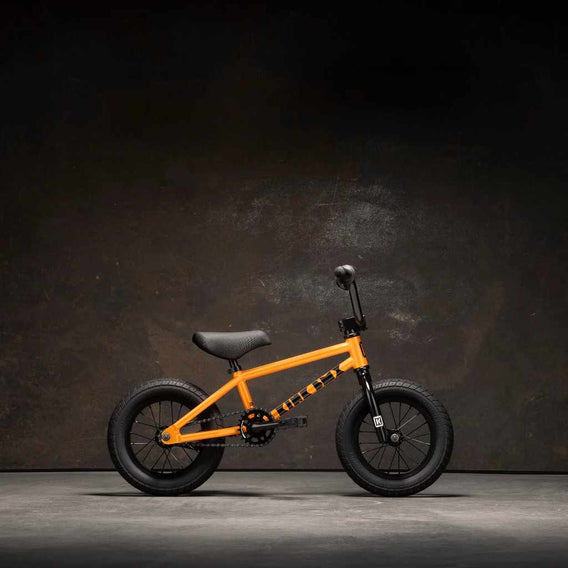 Kink 12" BMX Roaster complete bike in orange, side view photographed in an industrial warehouse