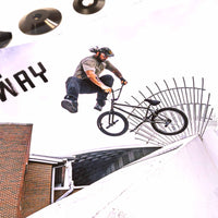 Person on a BMX bike performing a trick over a railing with a building and 'WAY' sign in the background.