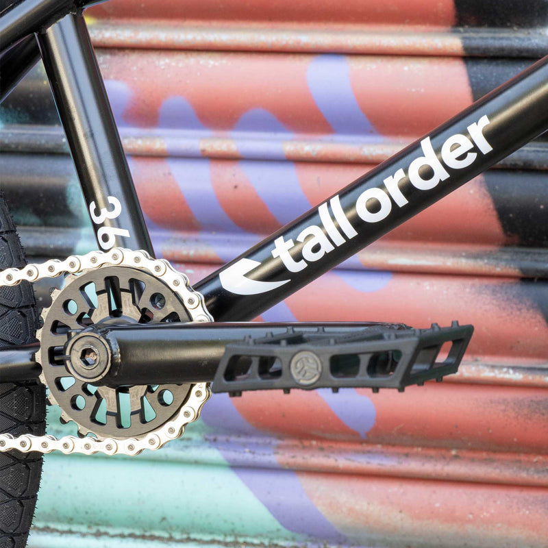 Close-up of a bicycle chain and gear system with 'tall order' branding against a colorful background.
